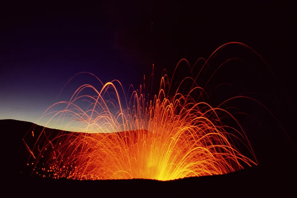 A volcano erupts at night, spewing glowing embers and lava trails into the dark sky.