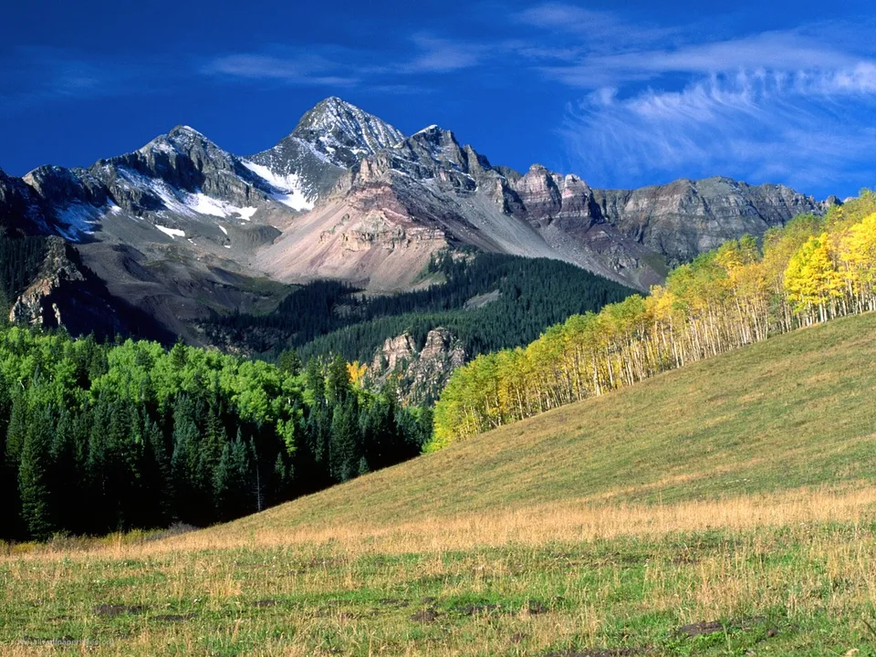 A majestic mountain range with snow-capped peaks, dense green forests, and a slope covered in golden aspen trees under a blue sky.