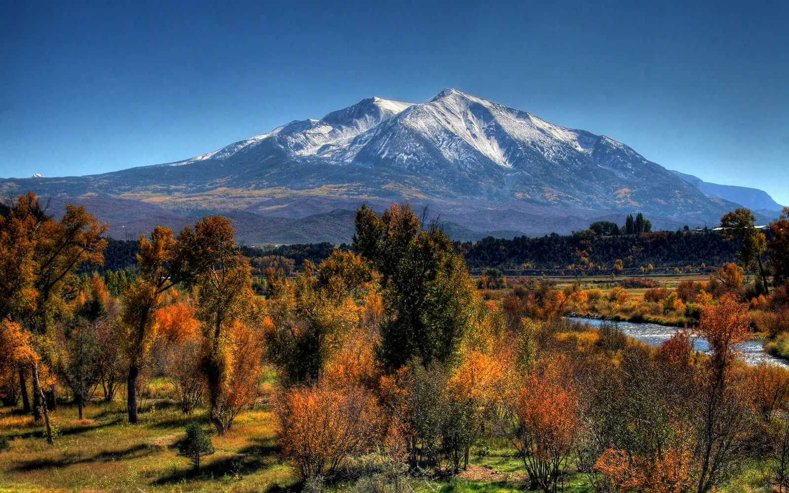 A snow-capped mountain looms over a valley filled with trees displaying vibrant autumn colors and a winding river.
