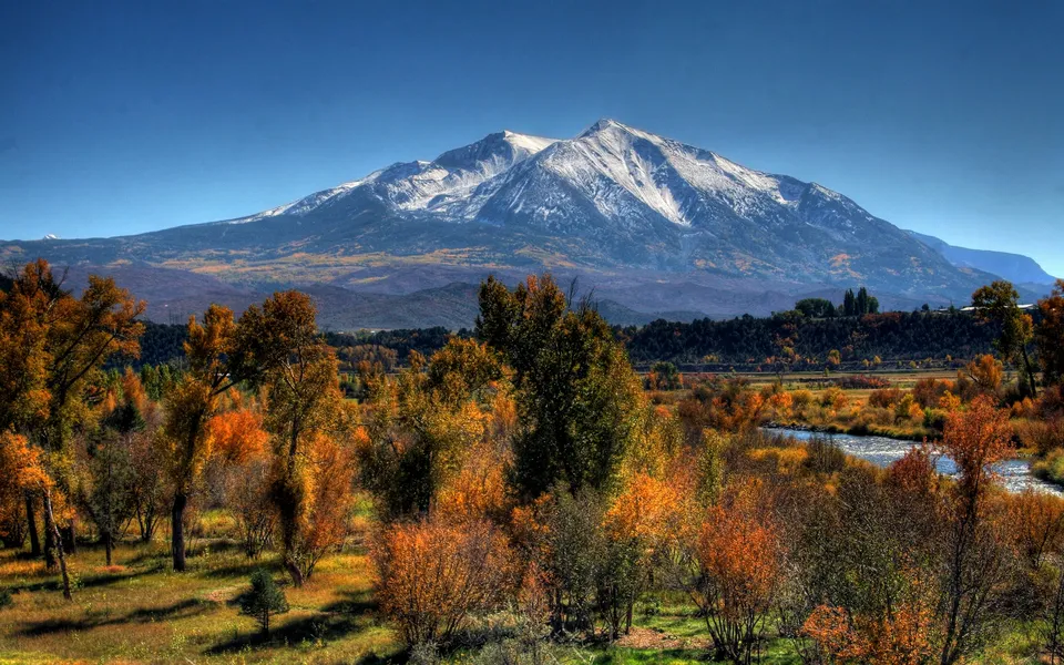 A snow-capped mountain looms over a valley filled with trees displaying vibrant autumn colors and a winding river.