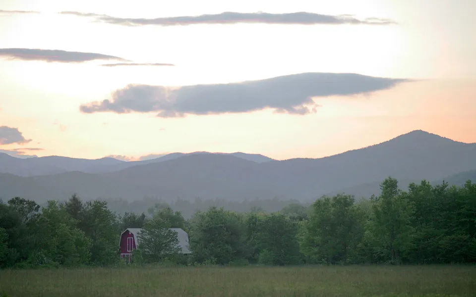 A red barn is nestled among trees with rolling mountains in the background under a soft, cloudy sky at dusk.