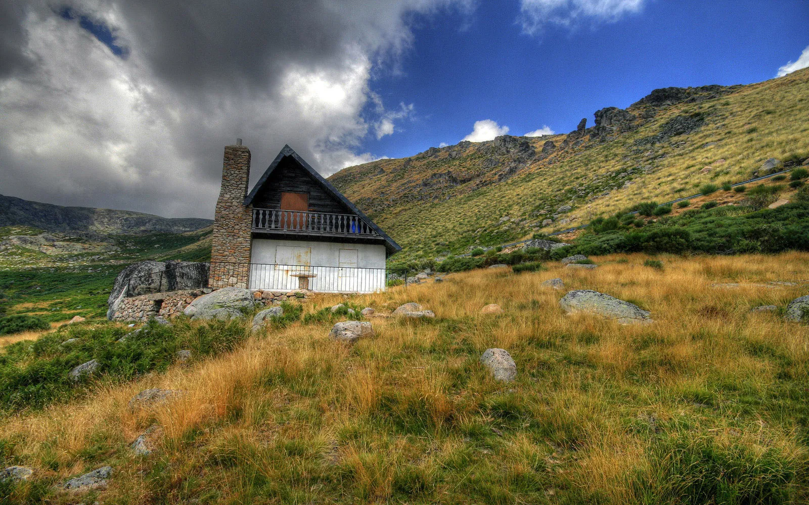 A rustic cabin with a stone chimney sits on a grassy mountain slope under a dramatic sky.