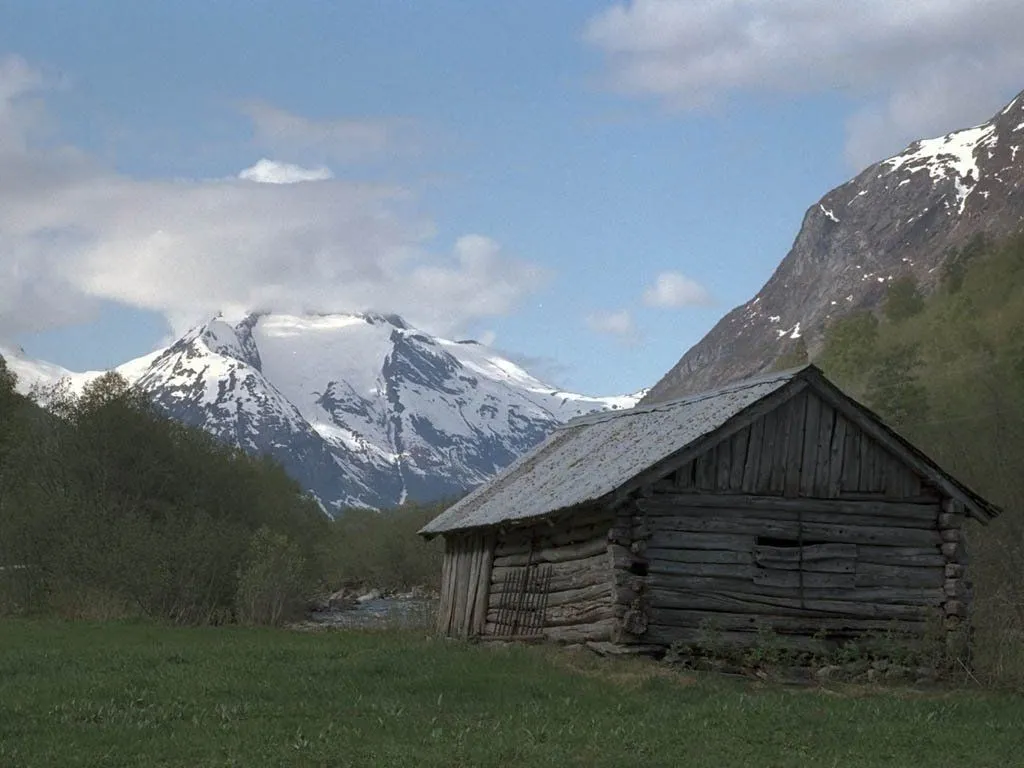 A weathered wooden cabin sits in a grassy field with a large snow-covered mountain range in the background.