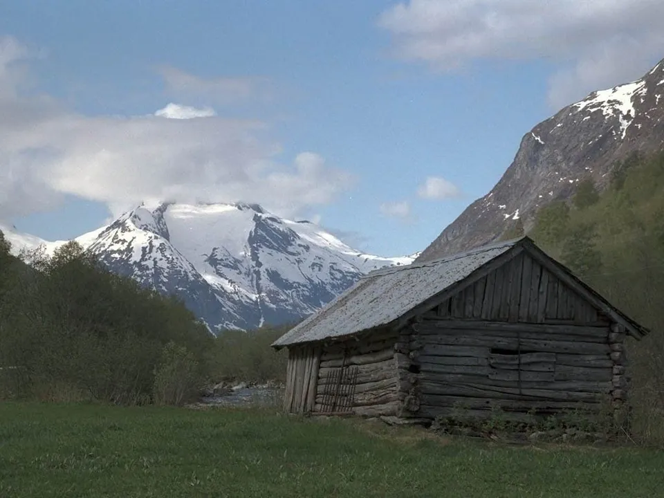 A weathered wooden cabin sits in a grassy field with a large snow-covered mountain range in the background.