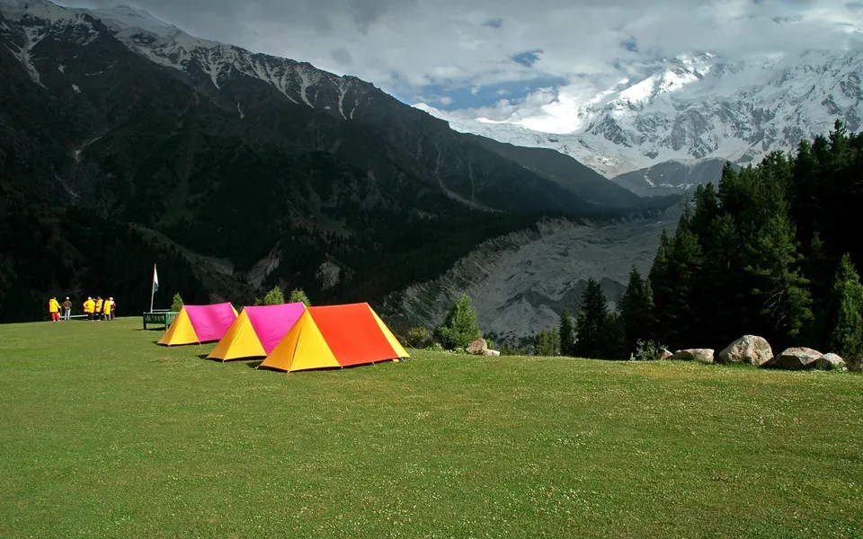 Several colorful camping tents are pitched on a vibrant green grassy meadow with snow-capped mountains in the background.