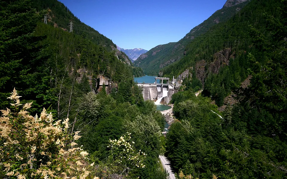 A wide shot of a concrete dam holding back a turquoise mountain lake, surrounded by lush green forests.