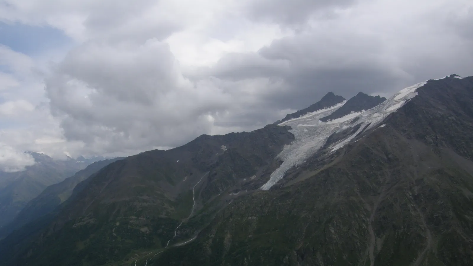 A vast mountain range with a prominent glacier flowing down its side under a cloudy sky.