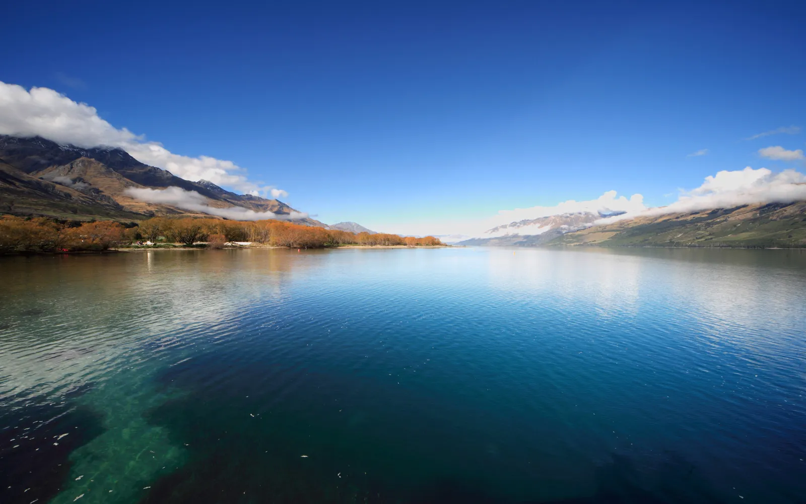 A wide, calm lake reflects the bright blue sky and distant mountains with scattered clouds.