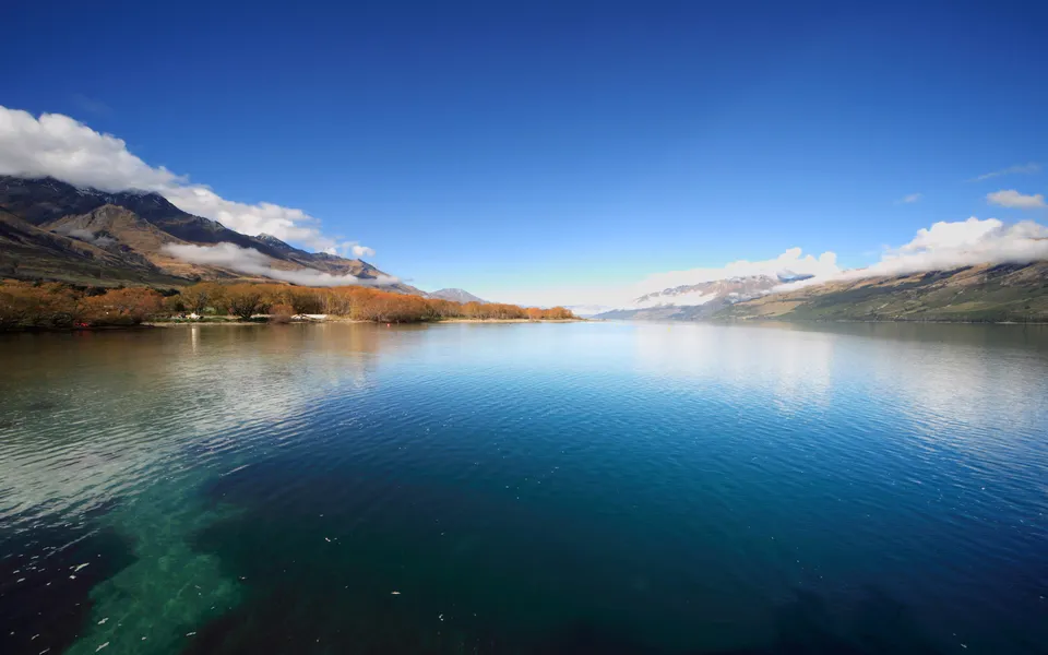 A wide, calm lake reflects the bright blue sky and distant mountains with scattered clouds.
