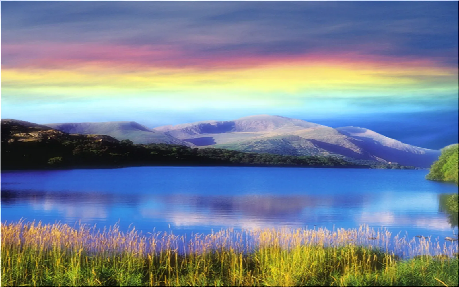 A wide, calm blue lake reflects a vibrant, multi-colored sky above rolling hills and mountains.