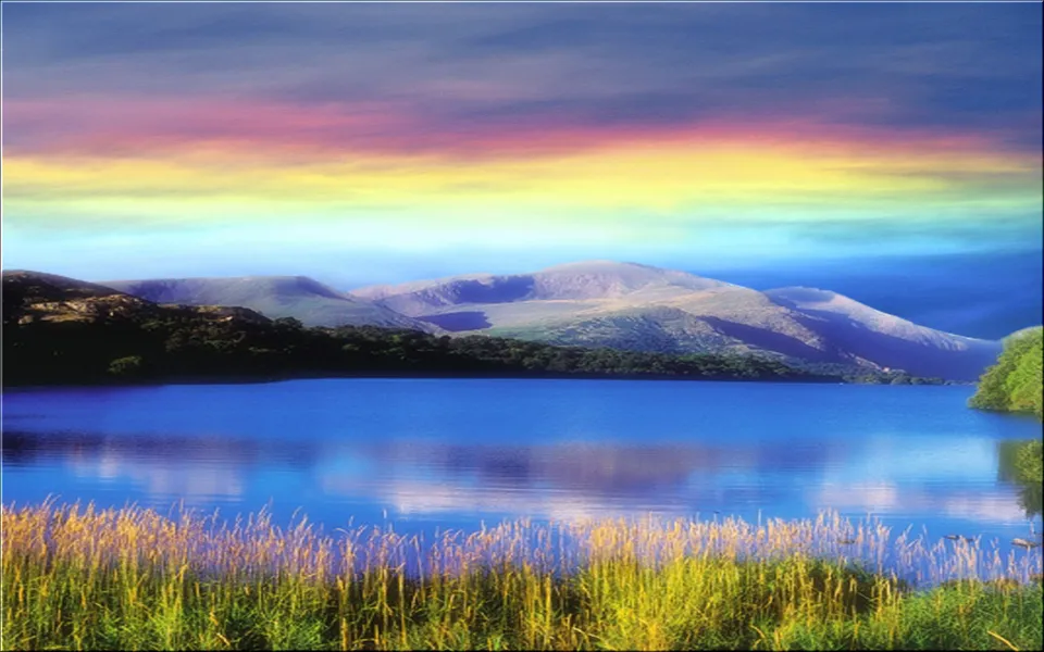 A wide, calm blue lake reflects a vibrant, multi-colored sky above rolling hills and mountains.