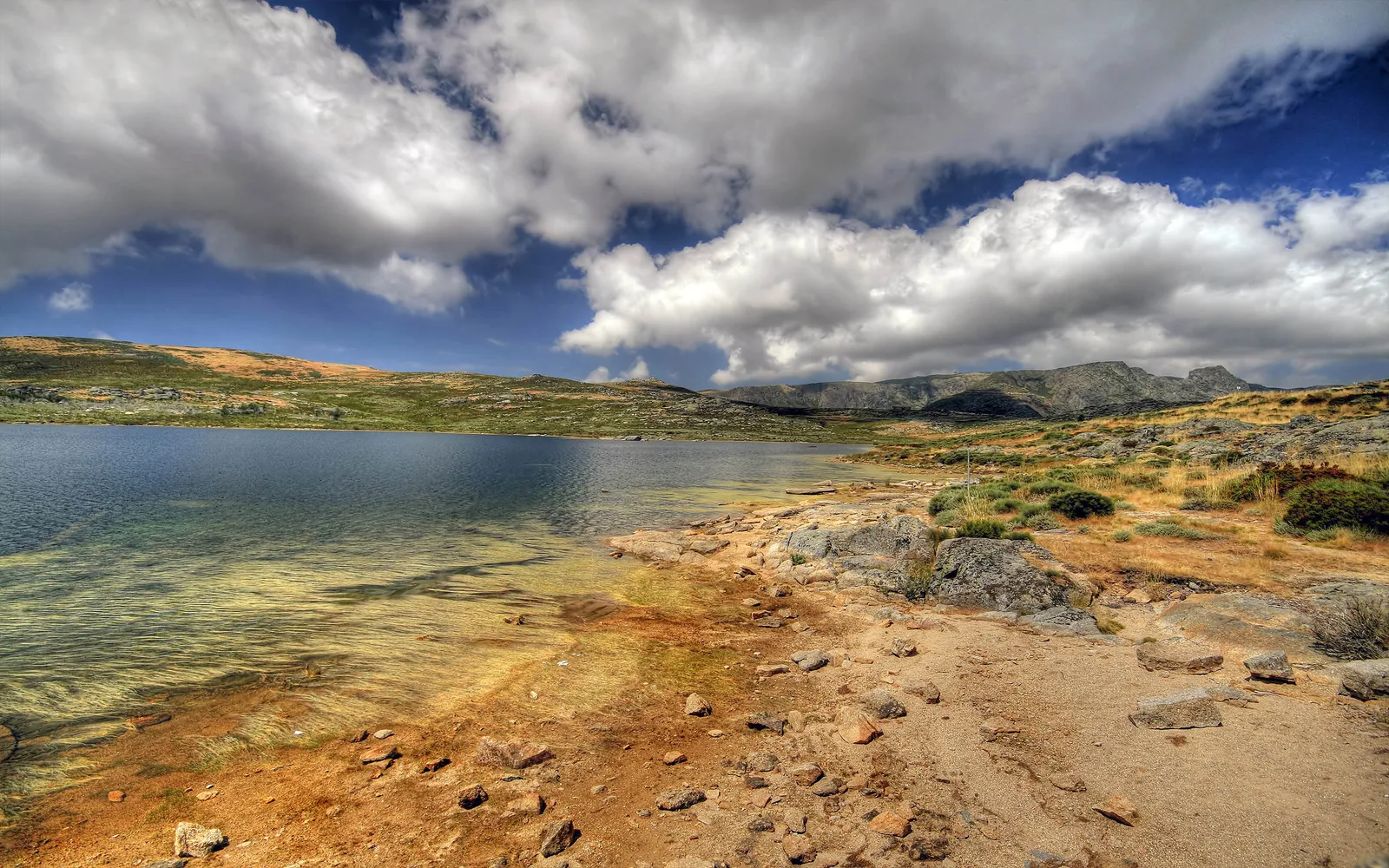 A wide shot of a calm lake with rocky shores and rolling hills under a sky filled with dramatic clouds.