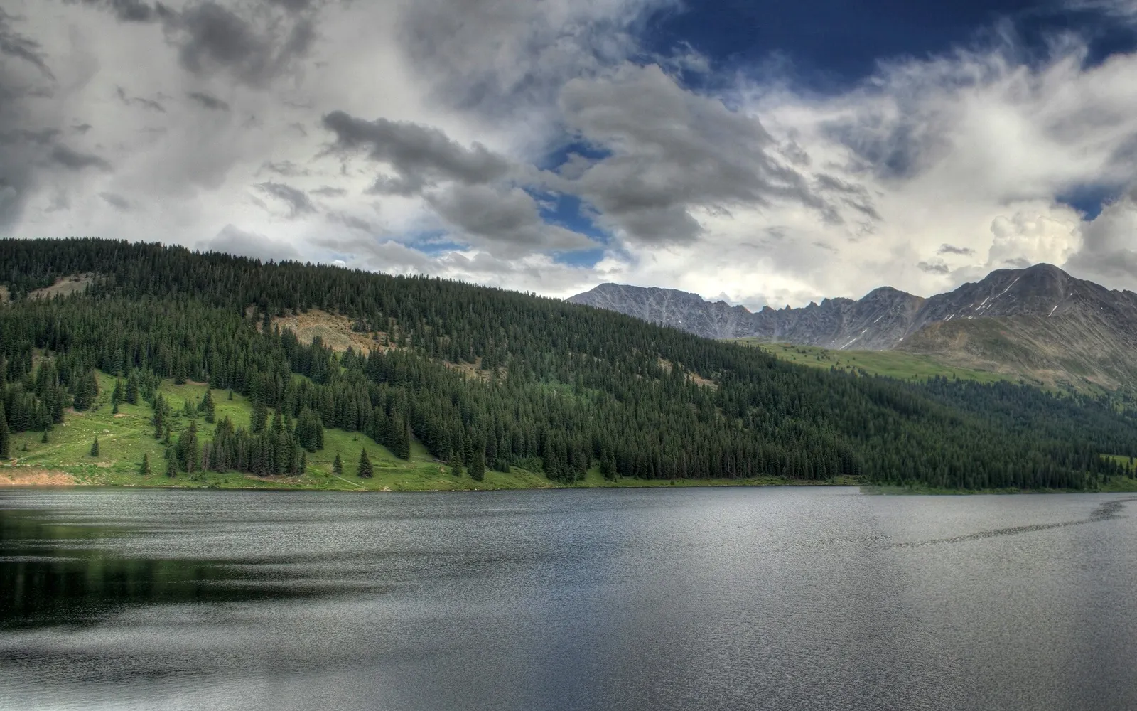 A wide, dark lake reflects the cloudy sky, with a densely forested mountain slope rising behind it.