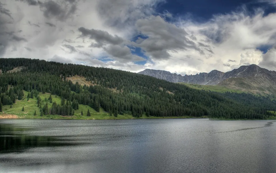 A wide, dark lake reflects the cloudy sky, with a densely forested mountain slope rising behind it.