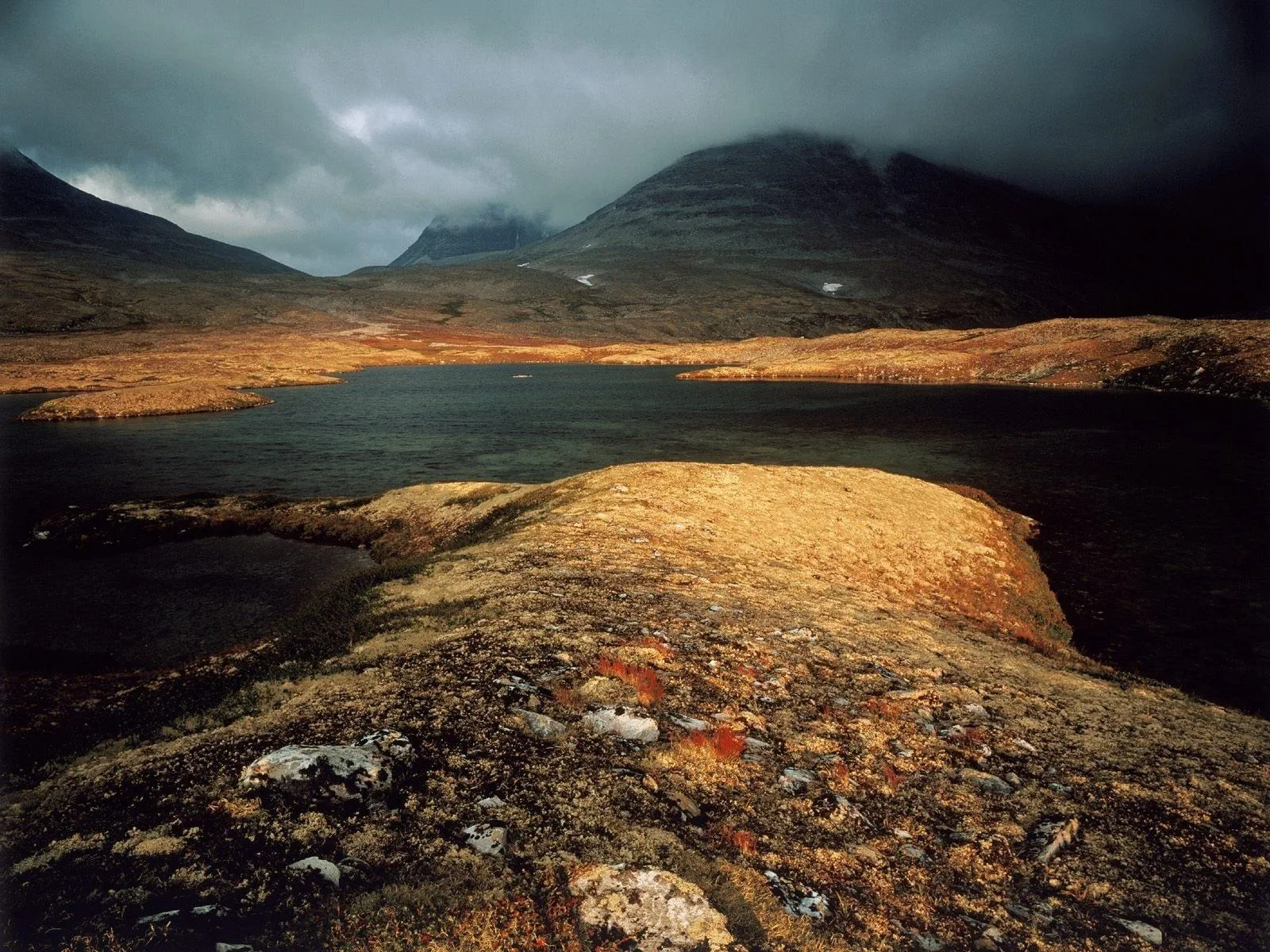 A dark lake is surrounded by hills covered in golden and brown vegetation, with dramatic clouds overhead.