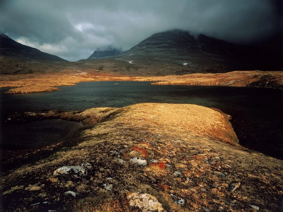 A dark lake is surrounded by hills covered in golden and brown vegetation, with dramatic clouds overhead.