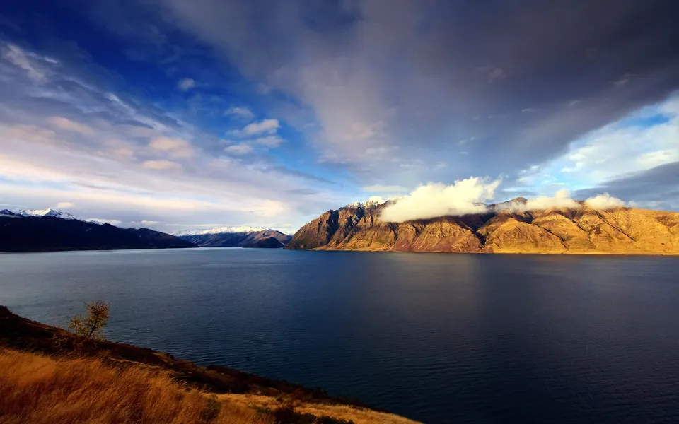 A wide, deep blue lake stretches towards distant snow-capped mountains under a dramatic sky with clouds.