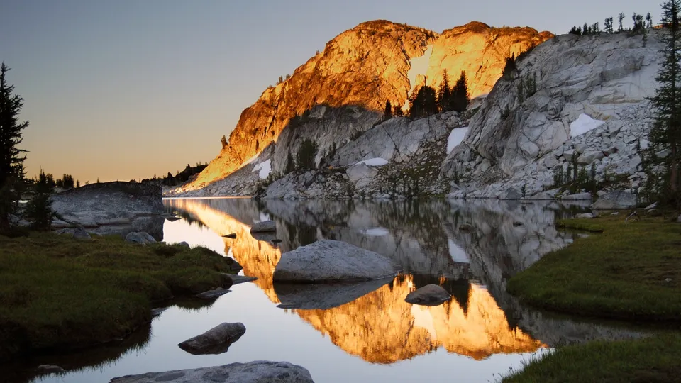 A mountain lake reflects the golden light of sunrise on a rocky peak, with green grass and trees in the foreground.