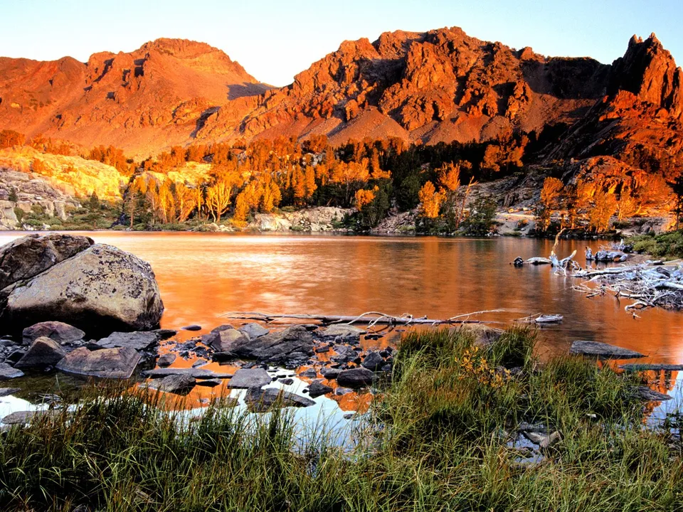 A serene mountain lake reflects the warm glow of sunset on rugged peaks and trees.