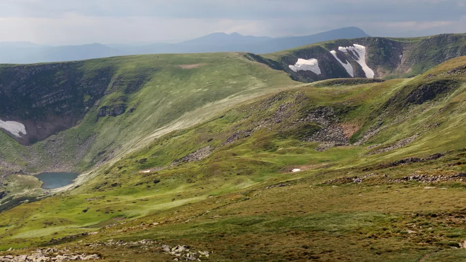 A small, dark blue lake sits in a valley between rolling green mountains with patches of snow.