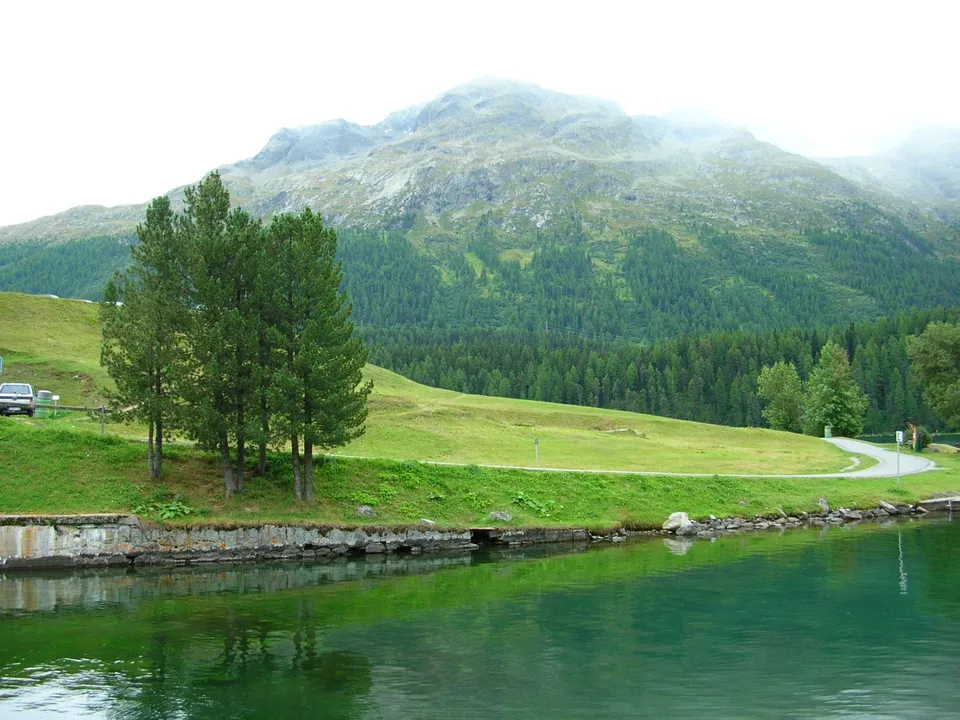 A calm, green lake reflects trees and a misty mountain range under an overcast sky.
