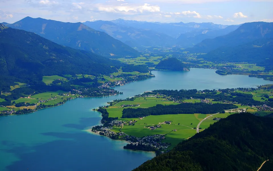 An aerial view of a vast turquoise lake surrounded by green hills and distant blue mountains under a partly cloudy sky.