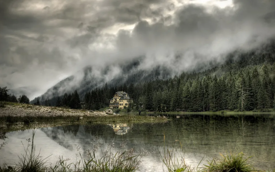 A yellow house sits on the shore of a lake, surrounded by pine trees and misty mountains.