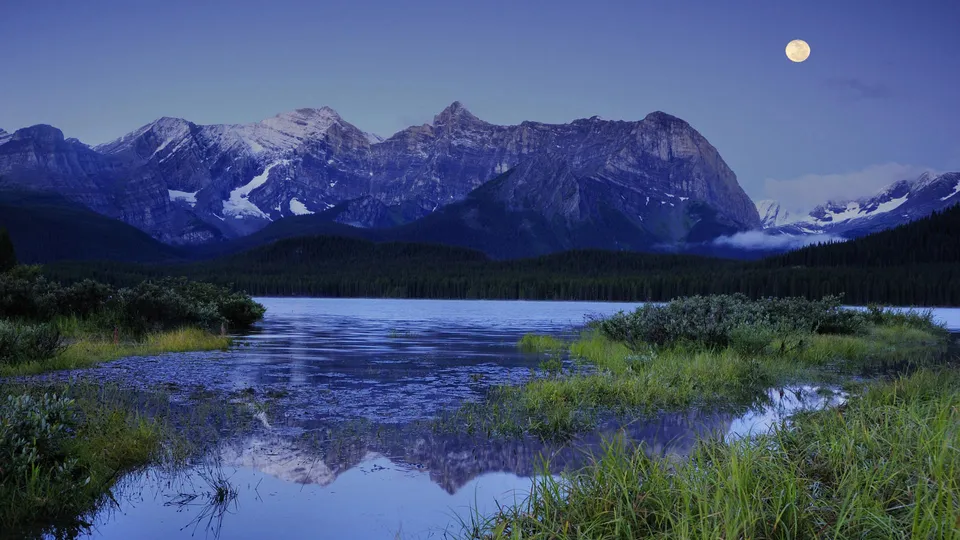 A tranquil lake reflects snow-capped mountains under a clear night sky with a full moon.