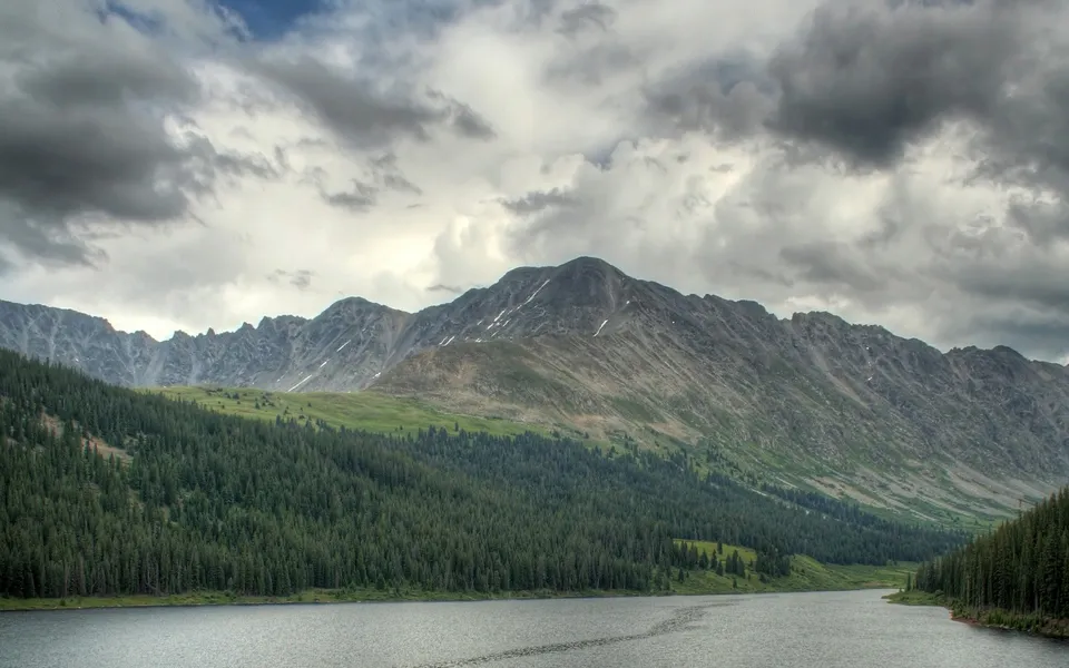 A wide, calm lake reflects the sky in front of a dense pine forest and rugged mountains under a cloudy sky.