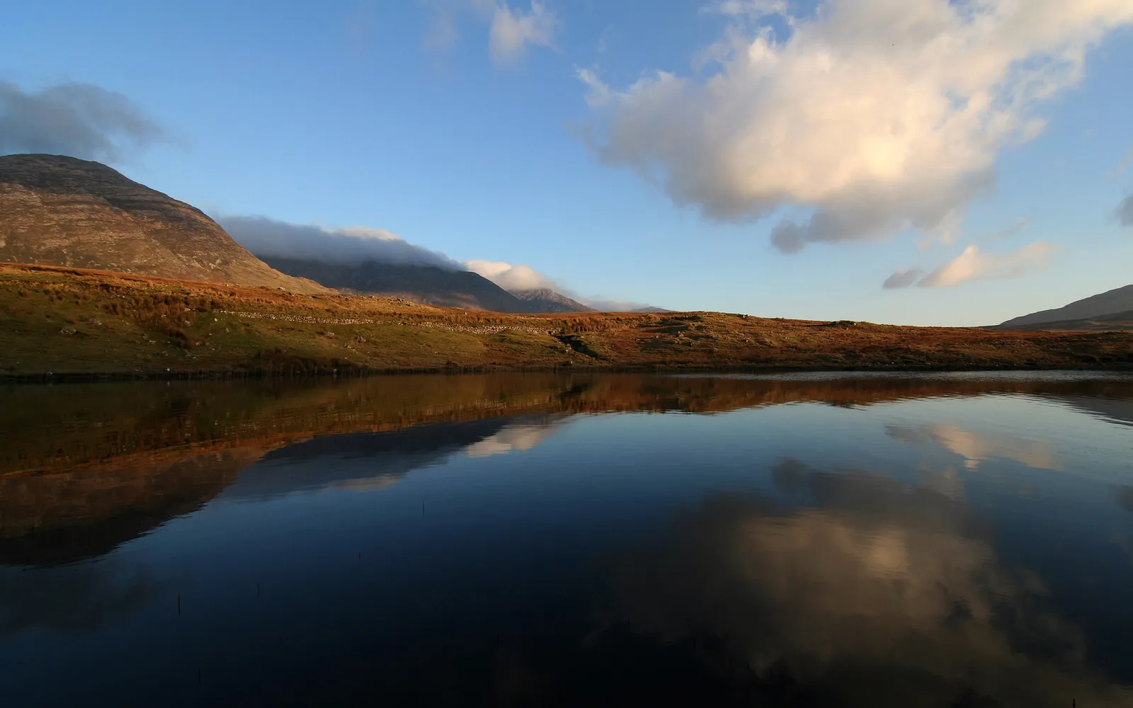 A calm lake reflects the sky, clouds, and a sloping mountain range under a clear blue sky.