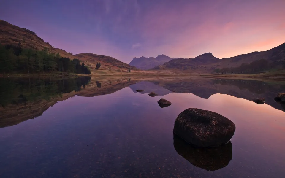 A calm lake reflects mountains and a colorful sky during sunrise, with rocks in the foreground.