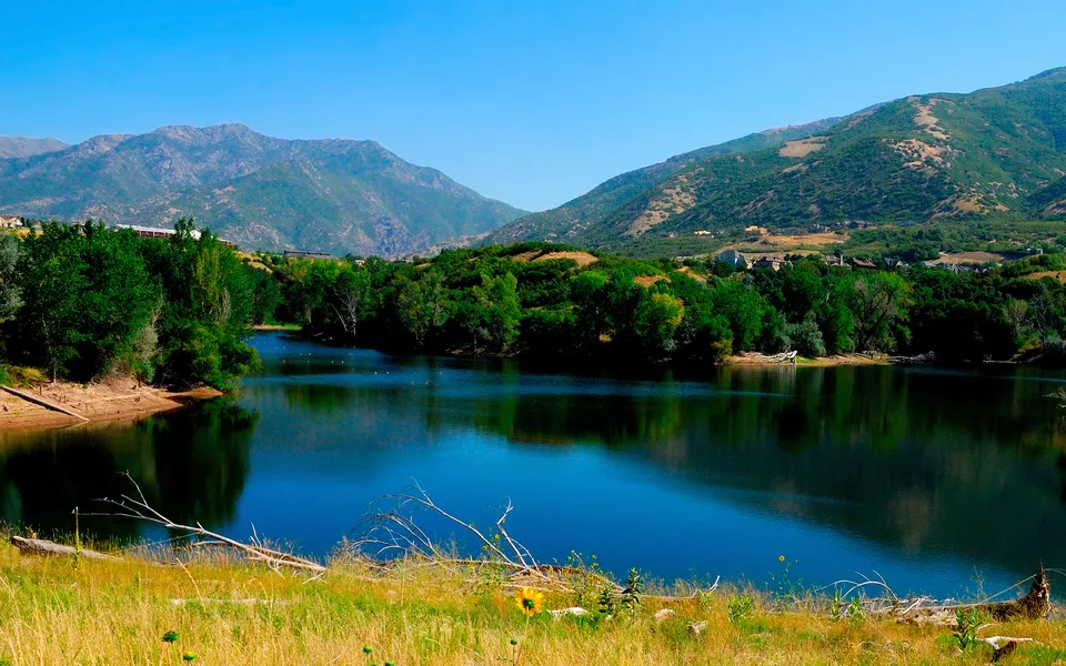 A calm, deep blue lake reflects the surrounding green trees and distant mountains under a clear blue sky.