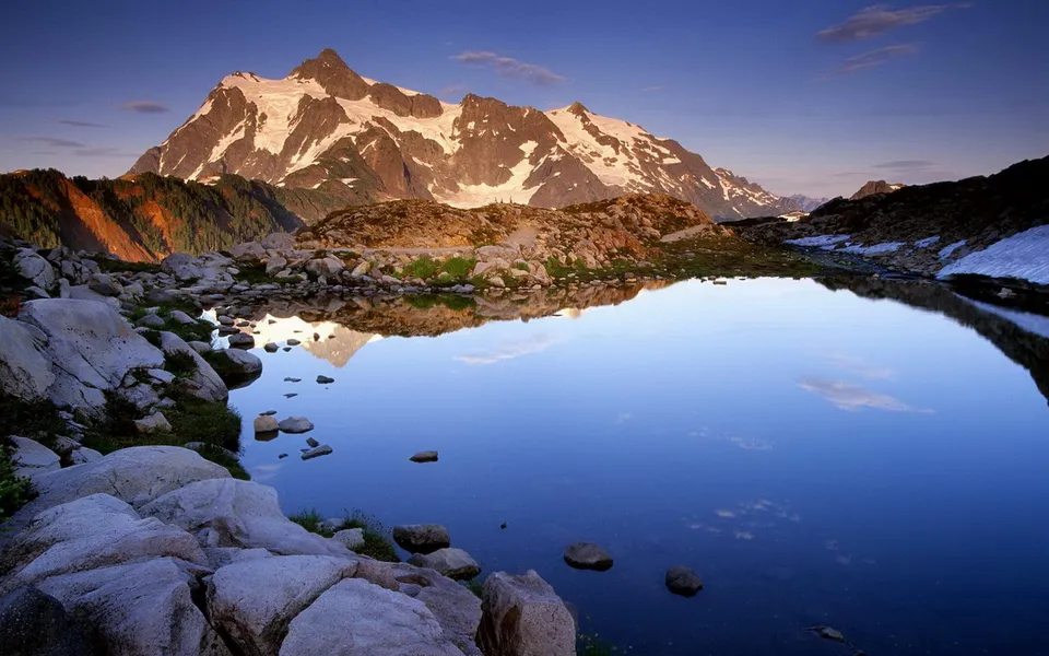 A snow-capped mountain range is reflected in a still, blue alpine lake, with rocky shores in the foreground.