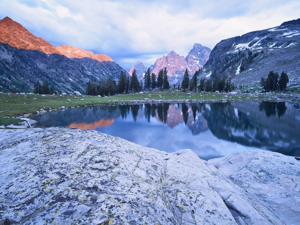 A calm mountain lake reflects the jagged, snow-dusted peaks and trees under a soft, cloudy sky.