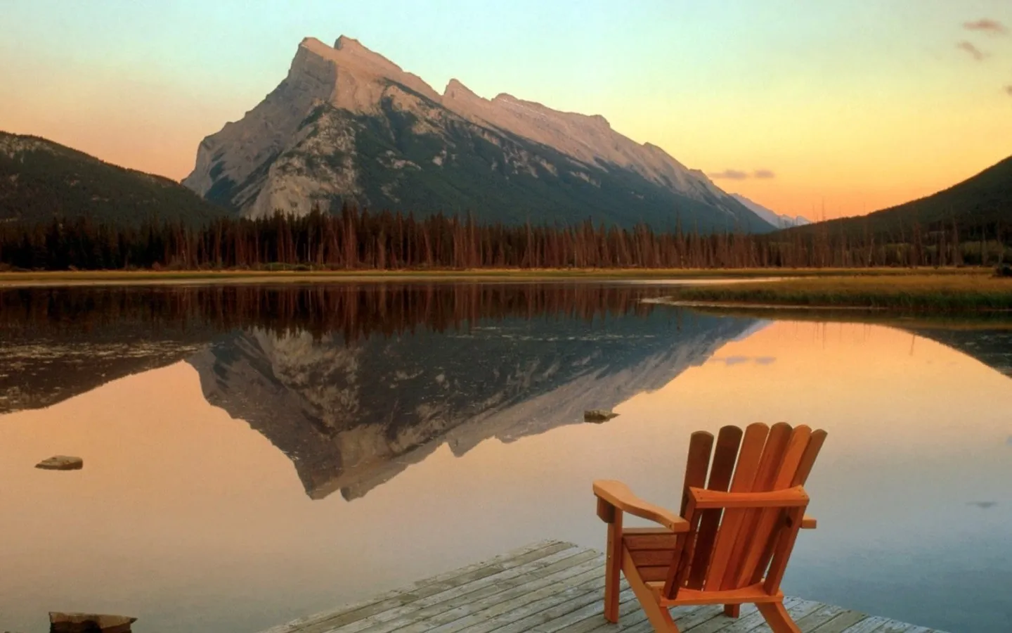 A wooden Adirondack chair sits on a dock facing a calm lake reflecting a majestic mountain and a colorful sunset sky.
