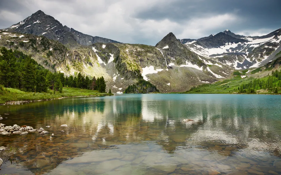 A clear turquoise mountain lake reflects snow-capped peaks and green trees under a cloudy sky.