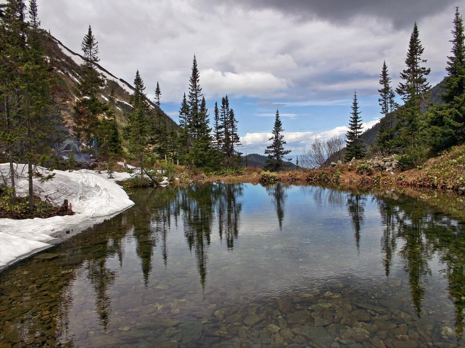 A clear mountain lake reflects evergreen trees and a cloudy sky, with patches of snow on the shore.