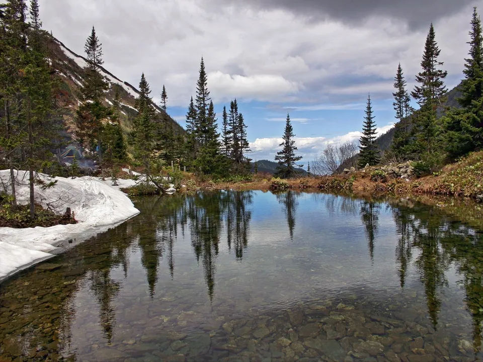 A clear mountain lake reflects evergreen trees and a cloudy sky, with patches of snow on the shore.