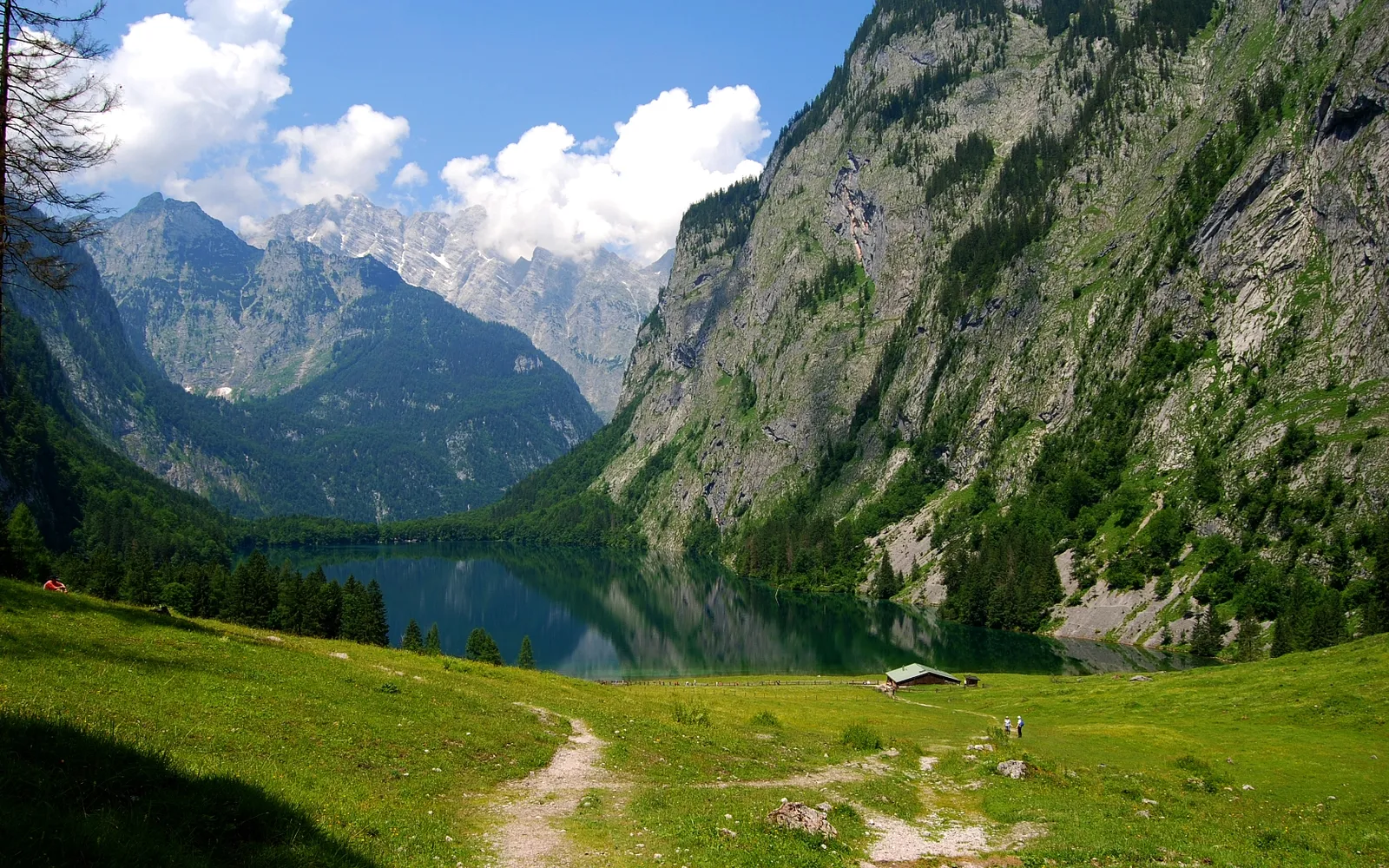 A tranquil lake reflects the surrounding steep, green mountains under a blue sky with white clouds.