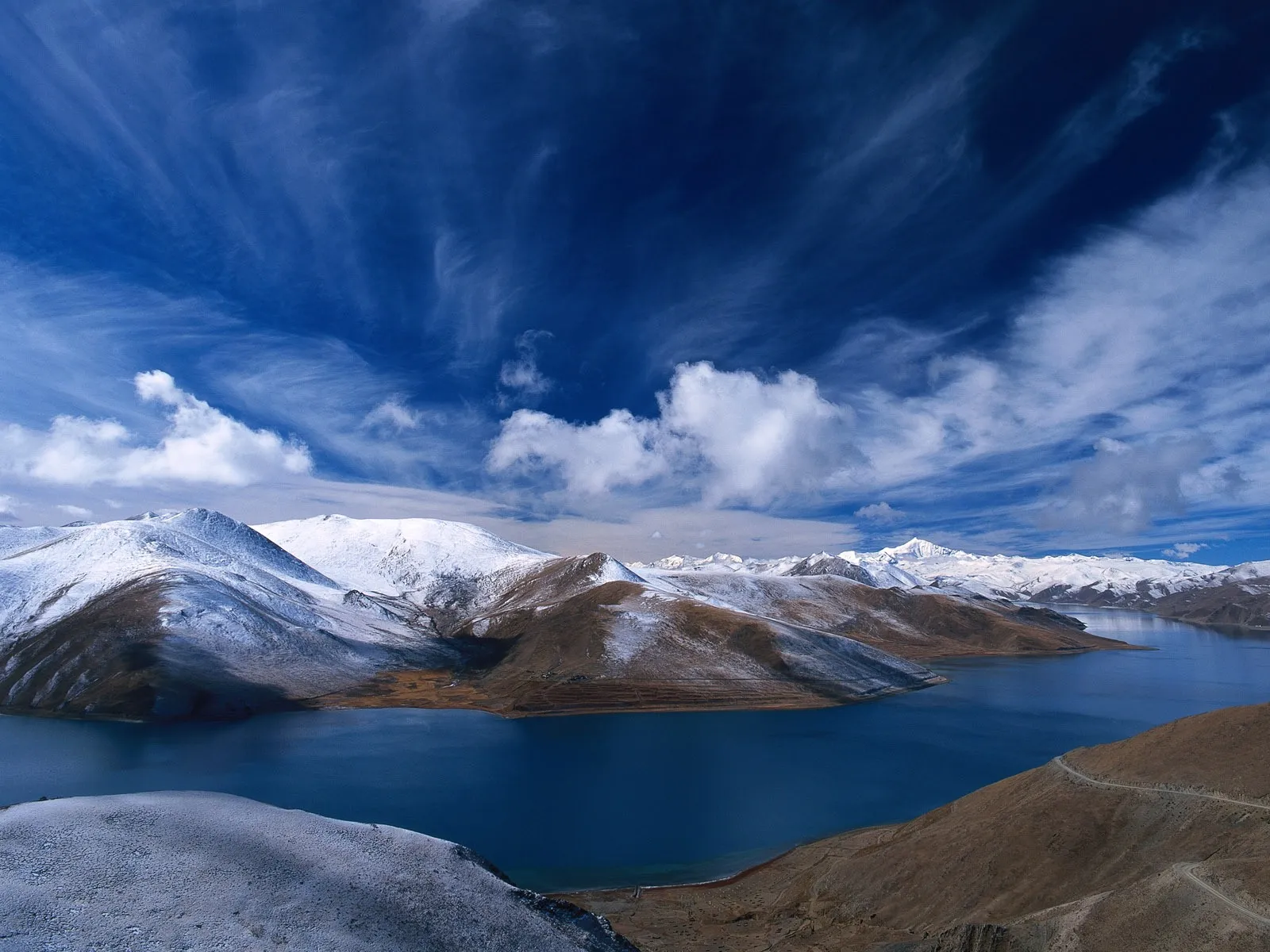 A wide, deep blue lake is nestled between rolling hills and majestic snow-covered mountains under a dramatic blue sky with wispy clouds.