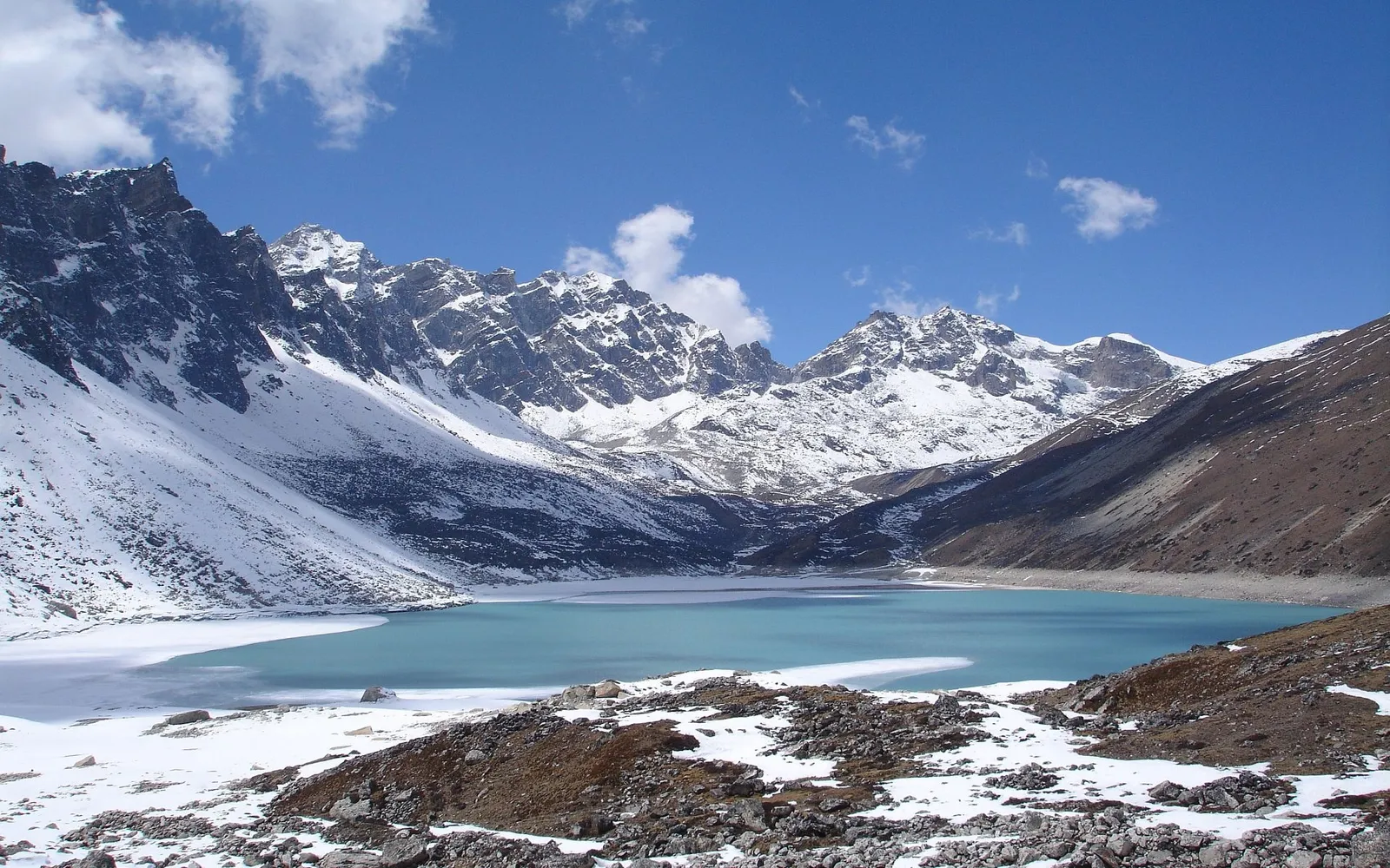 A bright turquoise lake is surrounded by snow-covered mountains and rocky terrain under a blue sky with clouds.