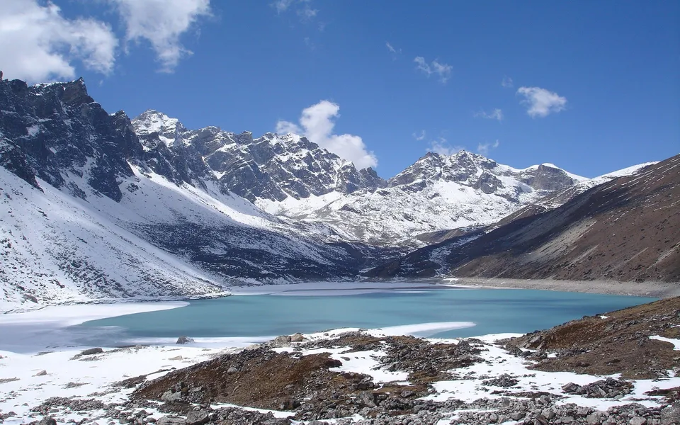 A bright turquoise lake is surrounded by snow-covered mountains and rocky terrain under a blue sky with clouds.