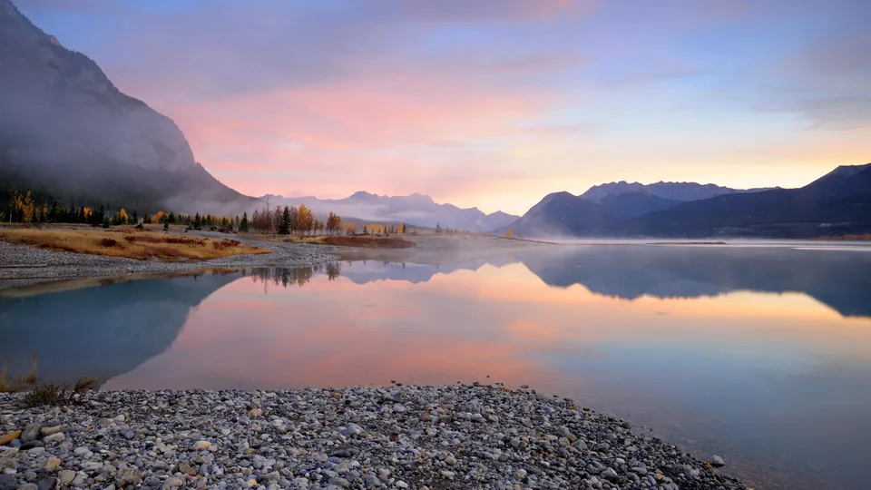 A serene mountain lake reflects the pastel colors of a sunrise sky, with mist rising from the water and mountains in the background.