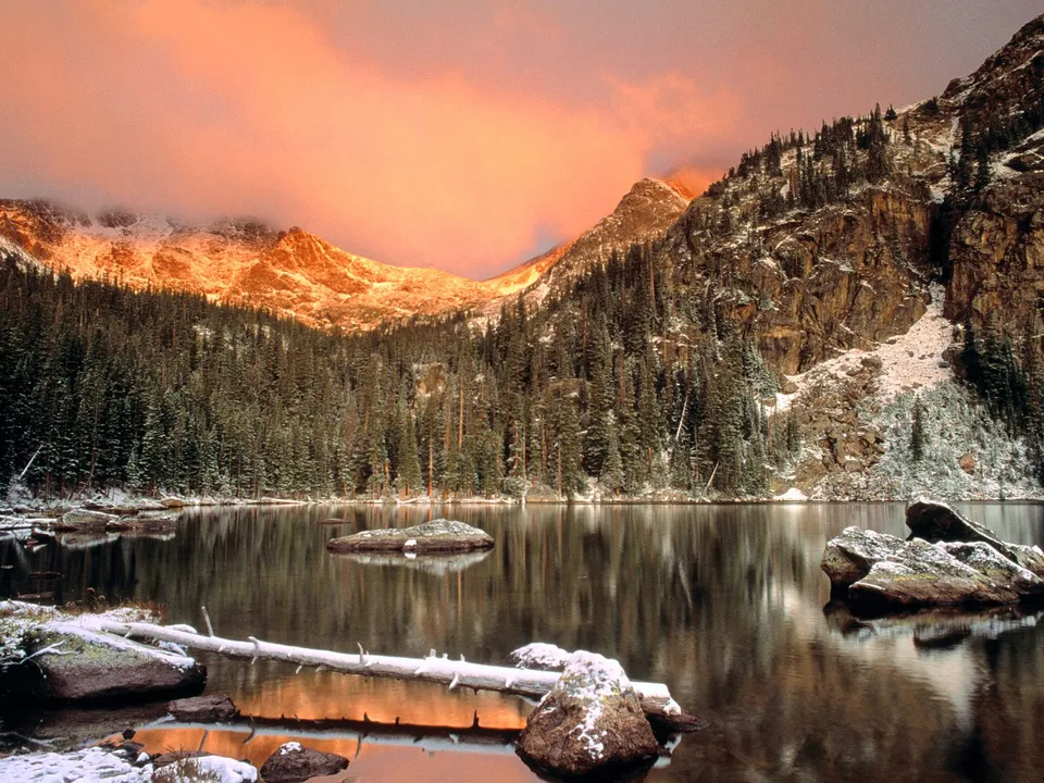 A serene mountain lake reflects snow-dusted peaks and a vibrant sunrise sky.
