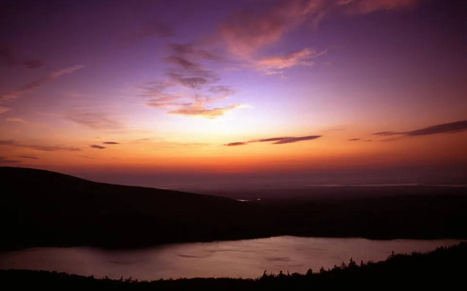A colorful sunrise paints the sky with purple and orange hues over a dark mountain silhouette and a calm lake.