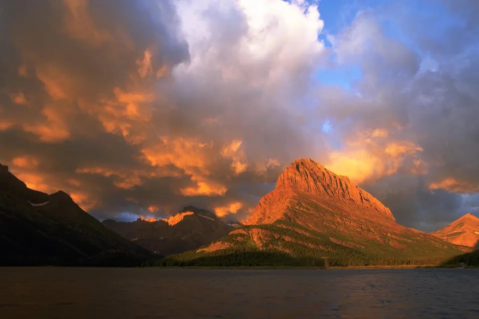 A mountain range is illuminated by the warm glow of a sunset, with dramatic clouds in the sky and a lake in the foreground.