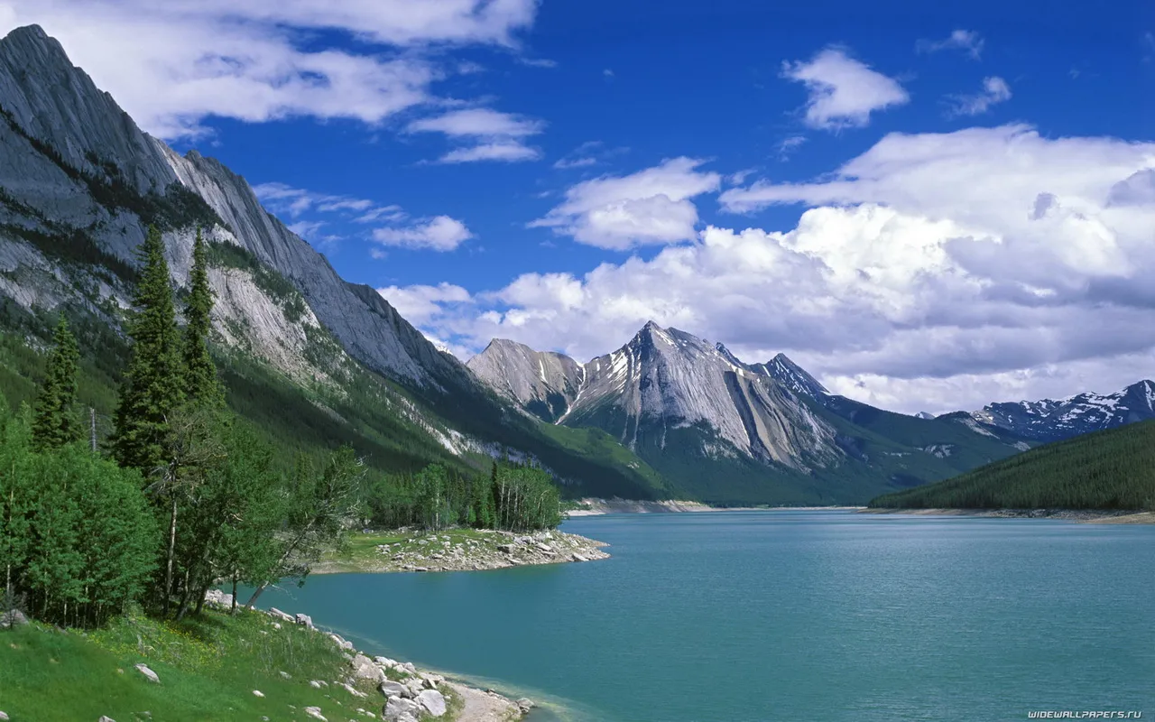 A wide, turquoise lake is bordered by green forested hills and dramatic, rocky mountains under a blue sky with white clouds.
