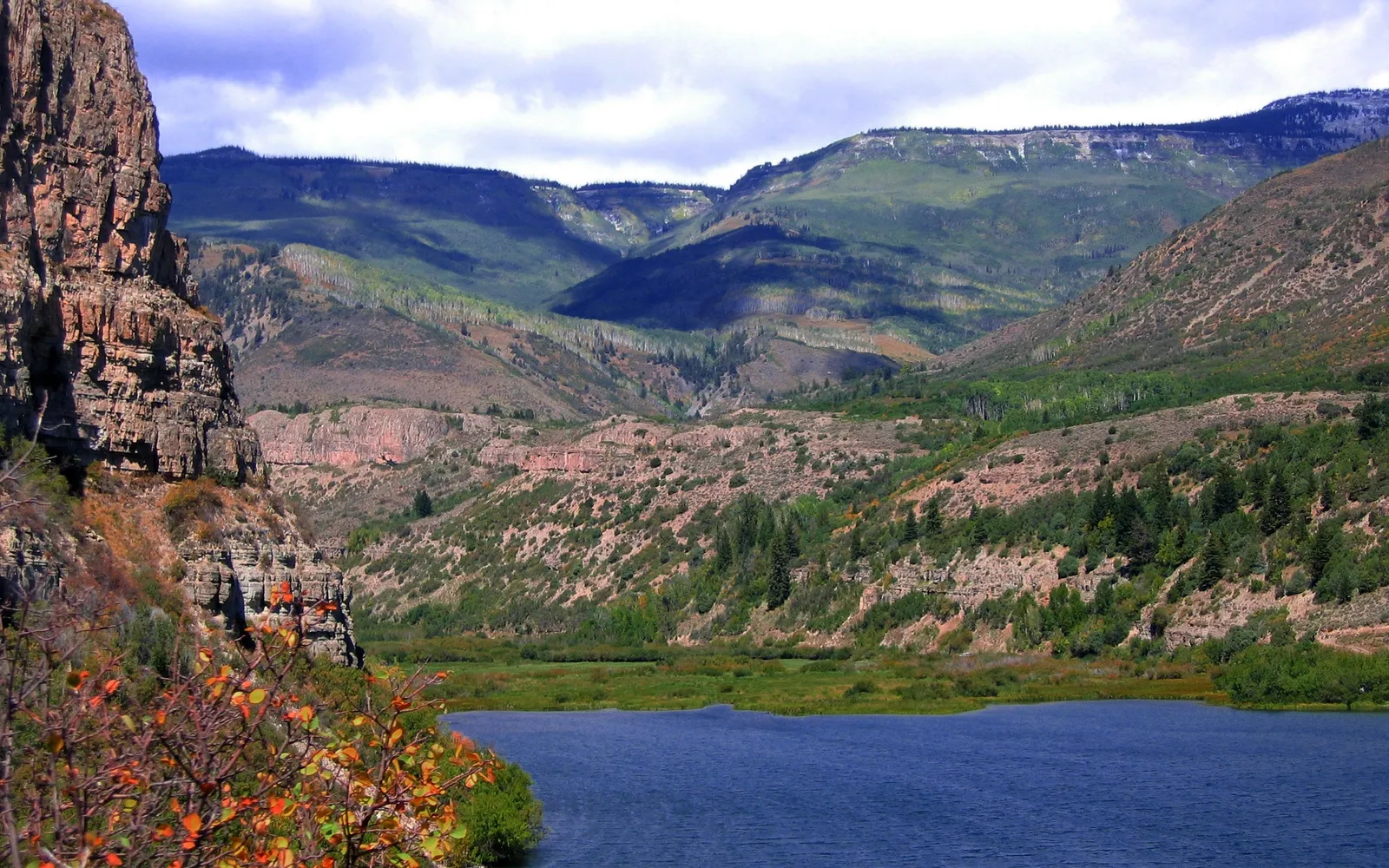 A calm blue lake is in the foreground, with rolling green hills and rocky cliffs in the background under a cloudy sky.