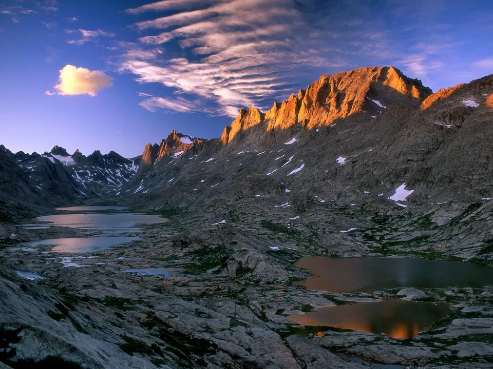A vast mountain range with jagged peaks, reflecting in calm alpine lakes under a dramatic sky.