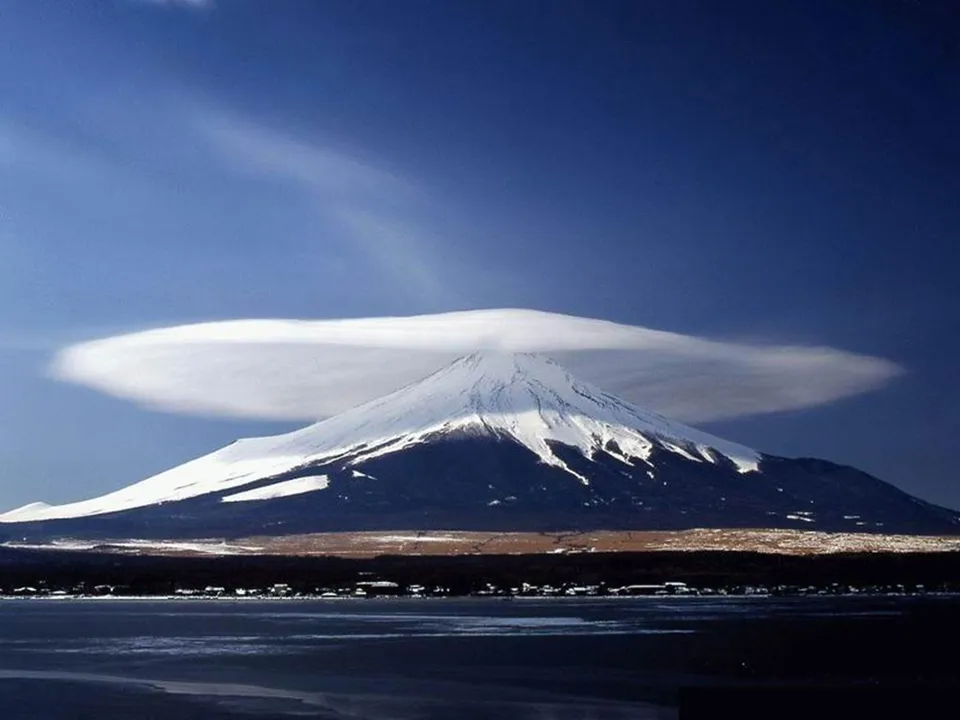 A snow-capped mountain peak is partially obscured by a large, flat, lenticular cloud.
